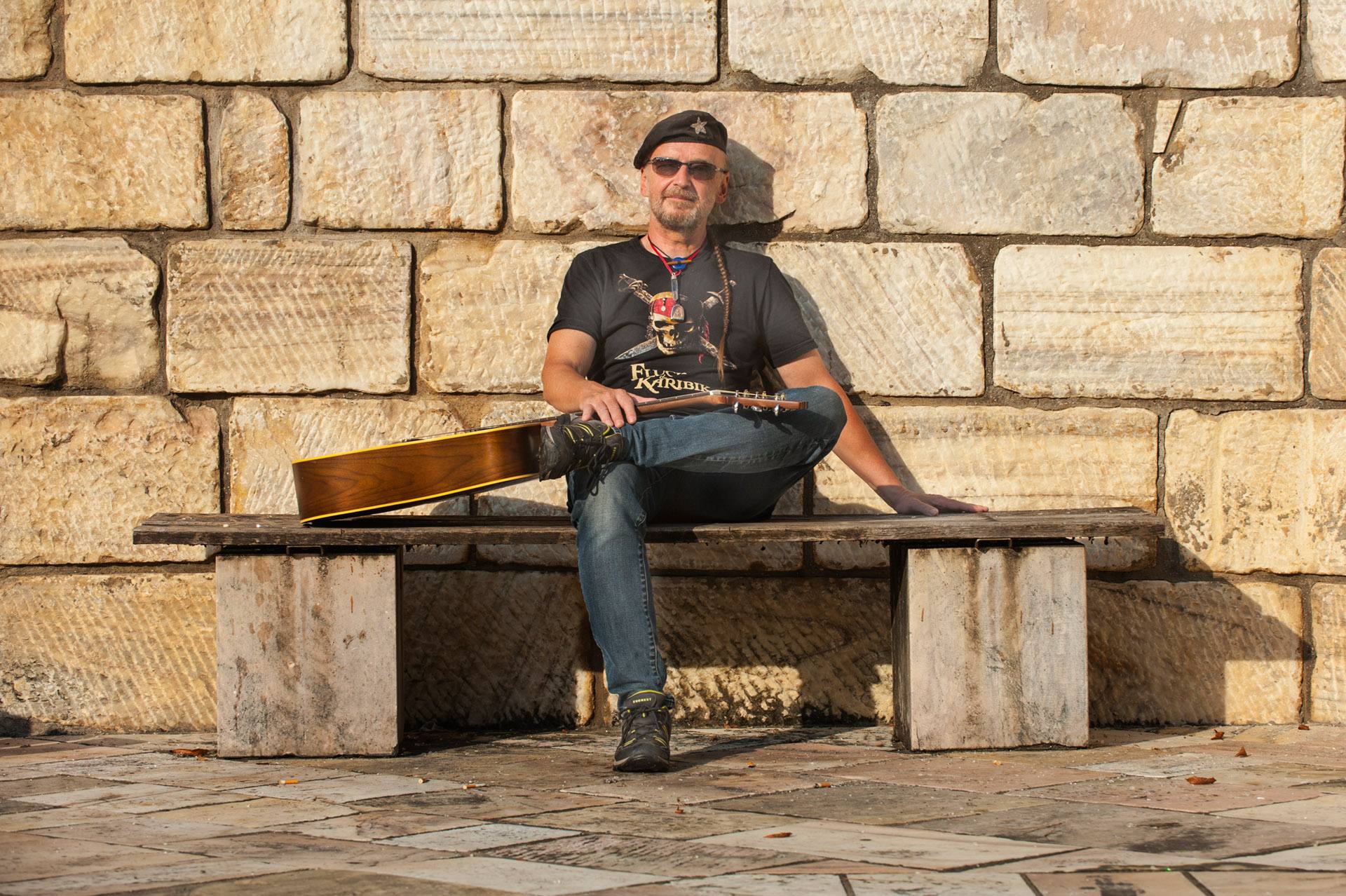 Thomas Goschat mit Gitarre auf einer Holzbank vor einer Sandsteinmauer